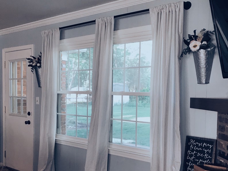 May include: Two windows with white striped curtains and a white door. The windows have a view of a green lawn and trees. There is a metal bucket with artificial flowers hanging on the wall to the right of the windows.