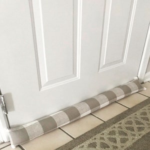 May include: A beige and white checked doorstop made of fabric. The doorstop is placed at the bottom of a white door.