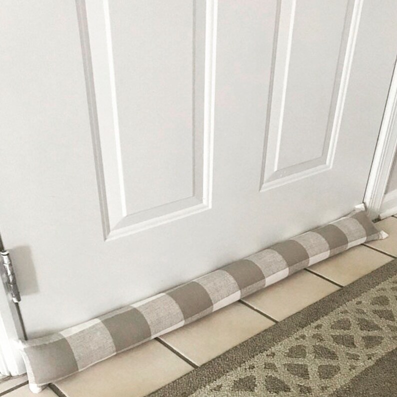 May include: A beige and white checked doorstop made of fabric. The doorstop is placed at the bottom of a white door.