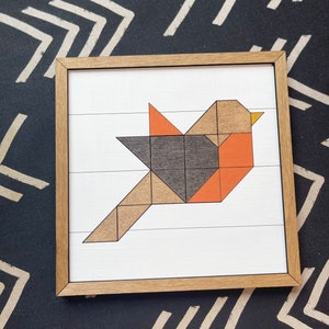May include: A framed geometric art print of a bird made from wood shapes in shades of brown, orange, and gray. The bird is flying with its wings spread out.