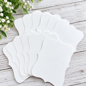 May include: A stack of white paper tags with a decorative scalloped edge and a stitched border detail. The tags are blank and rectangular with curved sides, arranged in a layered pile on a light-colored wooden surface.