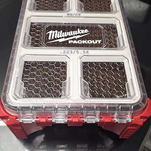 May include: A red and clear Milwaukee Packout storage organizer with five internal compartments filled with brass-cased ammunition. The lid features the text Milwaukee Packout, SG/CT, and .223/5.56, with a honeycomb pattern visible through the clear top.