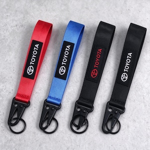 May include: Four fabric wrist lanyards with black metal clips and key rings. The lanyards come in red, blue, and two black versions. Each features a rectangular black patch with the white Toyota logo and text, except one black lanyard with red text.