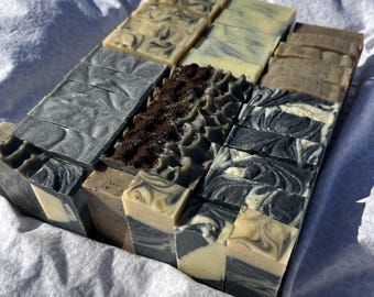 May include: A collection of rectangular soap bars arranged in rows. The bars feature various designs including solid cream, charcoal gray swirls, and one row topped with dark brown coffee grounds, showcasing a mix of textures and neutral tones.