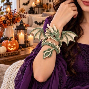 Könnte beinhalten: Ein gehäkeltes Drachenarmband in Salbeigrün und Creme. Der Drache hat detaillierte Flügel, einen langen, geringelten Schwanz und einen Kopf mit markanten Augen, das so gestaltet ist, dass es sich als Schmuckstück um das Handgelenk legt.