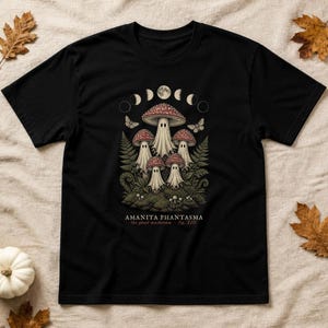May include: A black t-shirt featuring a graphic print of five ghost-like mushrooms with red caps and white bodies. Above the mushrooms are moon phases and butterflies. The text reads AMANITA PHANTASMA the ghost mushroom fig. XIII.