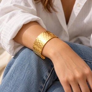 Puede incluir: Un brazalete ancho de tono dorado con una superficie martillada y texturizada. La banda de metal tiene un acabado pulido que refleja la luz sobre su exterior irregular y con hoyuelos, creando un patrón distintivo en toda la superficie de la joya.
