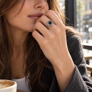Puede incluir: Un anillo de plata con una piedra negra redonda y pulida, montado en una banda de metal decorativa con intrincados patrones de remolinos y un borde de cuerda trenzada que rodea el engaste central.