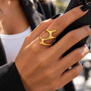 May include: A gold ring with an irregular, organic, open-work design featuring smooth, flowing lines and a polished metallic finish. The ring has a hollow, abstract shape that wraps around the finger, creating a distinct, sculptural appearance.