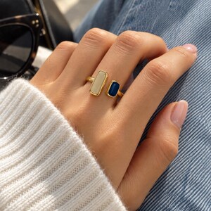 Puede incluir: Un anillo abierto de tono dorado con dos piedras rectangulares, una de color crema beige claro y la otra de color azul marino intenso, montadas en una banda de metal pulido que rodea el dedo con un espacio entre los dos engastes distintos.