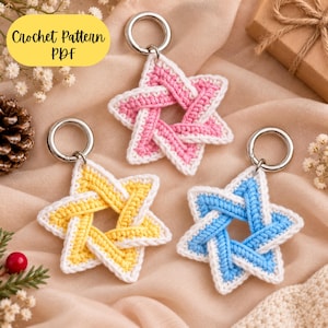 May include: Three crocheted star-shaped keychains with silver metal rings. Each star features a white border and a centre in either pink, yellow, or light blue. The text "Crochet Pattern PDF" is visible in the upper left corner of the image.