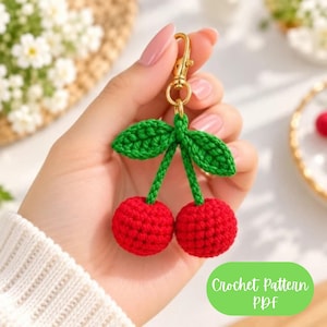 May include: A crochet cherry keychain featuring two round red spheres attached to green stems and two green leaves. The cherries are connected to a gold-colored metal lobster clasp. The image includes the text Crochet Pattern PDF.