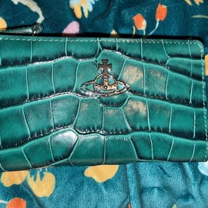 May include: A rectangular wallet in a deep emerald green hue featuring a glossy crocodile skin texture. Centred on the front is a polished silver-toned metal orb emblem with a cross on top. The wallet includes a zip closure along the top edge.