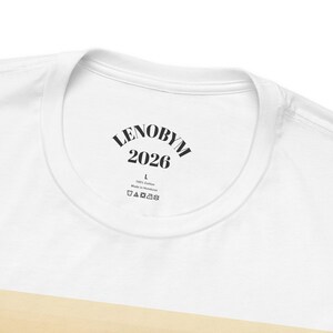 May include: A white t-shirt featuring a black printed inner neck label. The text reads LENOBYM 2026, followed by the size L, 100% Cotton, Made in Honduras, and a row of five standard garment care symbols including washing and drying instructions.