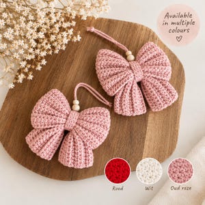 Op de afbeelding: Twee oudroze gehaakte strik-ornamenten, elk voorzien van een centrale houten kraal en een bijpassend ophanglusje. De tekst "Available in multiple colours" is zichtbaar, samen met kleine ronde stalen met de labels Rood, Wit en Oud roze.