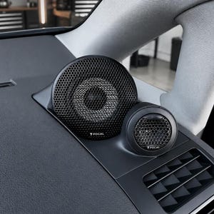 Może przedstawiać: Czarny samochodowy system głośników audio marki Focal zamontowany na desce rozdzielczej pojazdu. System składa się z większego okrągłego głośnika oraz mniejszego głośnika wysokotonowego, oba z czarną metalową kratką o strukturze plastra miodu.