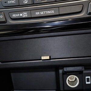 Op de afbeelding: Een zwart middenconsolepaneel van een voertuig met klimaatregelingsknoppen gelabeld SYNC, AUTO, REAR en RR SETTINGS. Daaronder bevinden zich een opbergvak, twee stoelverwarmingsknoppen, een ronde 12V-aansluiting en een USB-poort met 1,5A-icoon.