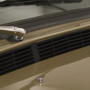 以下が含まれることがあります： タンカラーの車両のボンネット。黒い水平の換気グリル、金属製の銀色のワイパーアーム、そしてフロントガラスの近くの滑らかで光沢のある表面にある小さな円形の銀色の留め具が特徴です。