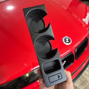 Peut inclure: Un insert de console centrale automobile en plastique noir mat doté de deux porte-gobelets circulaires et d'un compartiment de rangement rectangulaire avec un petit bouton circulaire à la base.