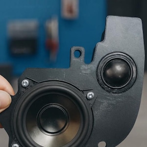 May include: A black automotive speaker assembly featuring a large circular woofer and a smaller circular tweeter mounted on a contoured plastic bracket. The unit is secured with four silver-colored metal screws and includes mounting holes.