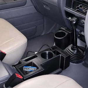 May include: A black plastic center console organizer installed between two beige car seats. The unit features two circular cup holders, a rectangular storage compartment containing a small blue item, and a gear shift lever nearby.