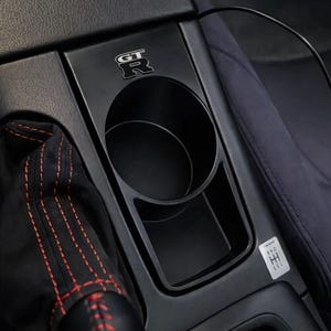 May include: A black automotive center console cup holder featuring a silver GT-R emblem. The unit includes a circular cup slot and a rectangular storage compartment, with a small gear shift pattern sticker labeled 1 3 5 and 2 4 R.