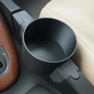 May include: A matte black cylindrical cup holder insert designed to fit into a vehicle center console. The holder features a smooth interior and a structured exterior base that integrates into the existing dark gray plastic console trim.