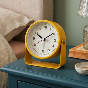 May include: A round yellow analog desk clock with a white face, black numerals, black hour and minute hands, and a thin red second hand. The clock is mounted on a matching yellow U-shaped metal stand that allows for a tilting motion.