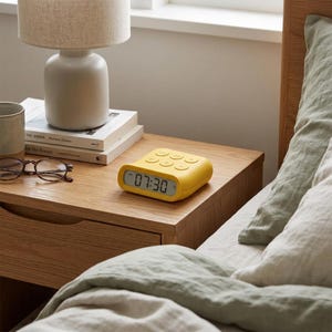 May include: A mustard yellow digital alarm clock with a rounded rectangular shape, featuring a front-facing LCD screen displaying the time 07:30 and six circular control buttons on the top surface for setting alarms and adjusting time settings.