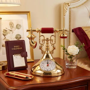 May include: A gold-toned rotary-style telephone with a red handle and a clock face integrated into the base. Next to it is a burgundy daily calendar, a gold pen on a small tray, and a white rose in a clear glass vase.