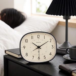 May include: An oval-shaped analog alarm clock with a cream-colored face, black numerals, and thin black hands. The clock features the text HoneyTime printed on the face. It has a black frame and sits on a flat surface, displaying a clear time-telling dial.