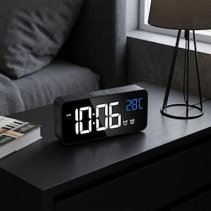 May include: A rectangular black digital alarm clock with a mirrored front display showing the time 10:06 AM in large white digits and the temperature 28 degrees Celsius in blue digits, featuring two alarm icons on the bottom right corner.