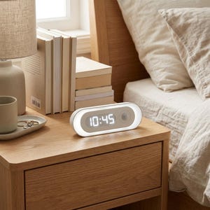 May include: A white, oval-shaped digital alarm clock with a glowing light border and a dark circular sensor on the right side. The clock face displays the time 10:45 in bright white digital numerals, positioned on a wooden nightstand surface.