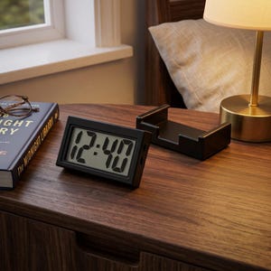 May include: A rectangular black digital alarm clock with a light gray screen displaying the time 12:40 in bold black digits. The clock features a simple frame and is positioned on a dark wood surface next to a black stand and a book.
