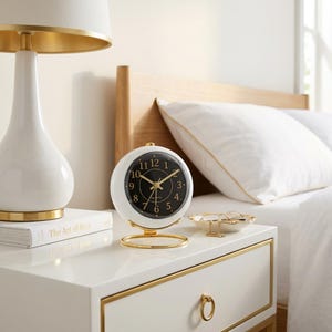 May include: A round analog table clock with a white outer casing and a black face featuring gold-colored numerals and hands. The clock is supported by a thin, circular gold-toned metal stand and includes the word Quartz printed on the clock face.