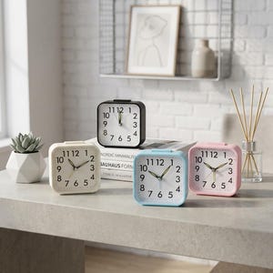 Könnte beinhalten: Vier quadratische analoge Wecker in Weiß, Schwarz, Hellblau und Pink. Jede Uhr hat ein weißes Zifferblatt mit schwarzen arabischen Ziffern, dünnen Stunden- und Minutenzeigern sowie einem Sekundenzeiger. Die Uhren stehen auf einer Fläche.