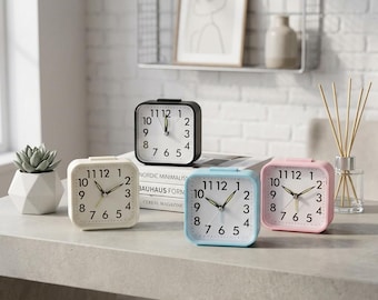 Puede incluir: Cuatro despertadores analógicos cuadrados en blanco, negro, azul claro y rosa. Cada reloj presenta una esfera blanca con números arábigos negros, manecillas finas para las horas y los minutos, y un segundero. Los relojes están dispuestos.