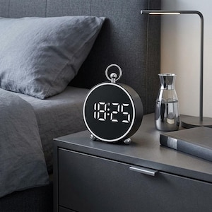 May include: A round digital alarm clock with a matte black finish and a silver ring handle on top. The clock face features a black background with bright white digital numbers displaying 18:25, surrounded by a thin, illuminated white circular border.