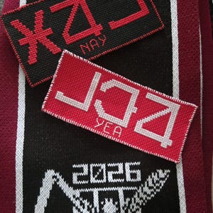 Könnte beinhalten: Ein schwarz-kastanienbrauner Strickschal mit weißen vertikalen Streifen und einer weißen Grafik einer Figur mit dem Text 2026. Darauf liegen zwei rechteckige bestickte Aufnäher: einer in Schwarz mit rotem Text NAY und einer in Rot mit weißem Text YEA.