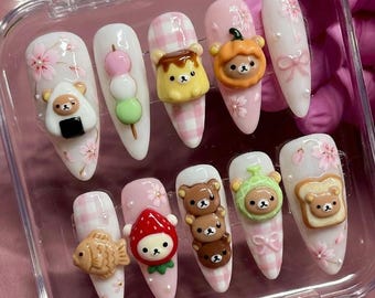 May include: A set of ten press-on nails featuring 3D charms of bears, food items like dango, taiyaki, and toast, and decorative elements including pink gingham patterns, cherry blossoms, and bows on white and light pink backgrounds.