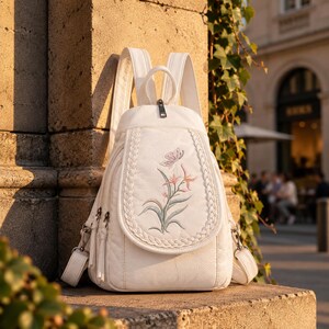 Puede incluir: Una mochila blanca con una solapa frontal con borde trenzado y un diseño bordado de una mariposa y flores rosas. La bolsa incluye correas ajustables para los hombros, un asa superior y herrajes de metal plateado con cierres de cremallera.