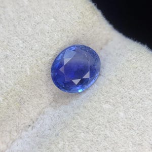 Op de afbeelding: Een gefacetteerde, ovaalvormige blauwe saffier edelsteen met een briljant slijpvorm, die diep koningsblauwe tinten en heldere interne reflecties vertoont, rustend op een gestructureerd, gebroken wit stoffen oppervlak met een zachte vezelige textuur.