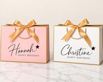 Pode incluir: Duas sacolas de presente retangulares, uma rosa claro e uma branca, com bordas douradas, laços de fita dourada e alças de corda dourada. A sacola rosa exibe o nome Hannah e a branca o nome Christine, ambas com o texto HAPPY BIRTHDAY e uma estrela preta.