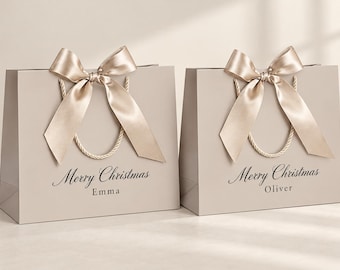 May include: Two light beige gift bags with twisted rope handles and large satin bows. Each bag features the black printed text Merry Christmas followed by a name, Emma on the left bag and Oliver on the right bag, in a serif font.
