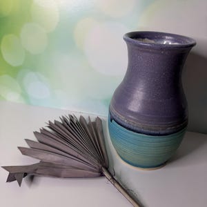 May include: A ceramic vase with a speckled purple upper section and a ribbed teal lower section, accompanied by a dried, fan-shaped palm leaf in a muted grey tone. The vase features a flared rim and a rounded body with a smooth finish.