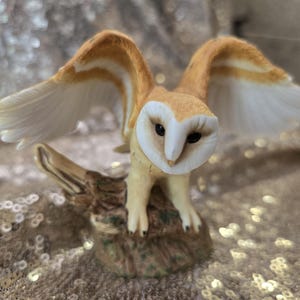 May include: A barn owl figurine with outstretched wings, featuring a white heart-shaped facial disc, dark eyes, and tan-colored plumage. The owl is perched on a textured, brown, bark-like base, capturing the bird in a mid-flight pose.
