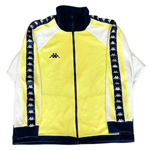 Op de afbeelding: Een geel en wit trainingsjack met een hoge kraag en een ritssluiting over de hele lengte. De mouwen hebben een donkerblauwe streep over de lengte met een herhalend wit Kappa-logopatroon. Een donkerblauw Kappa-logo staat op de rechterborst.
