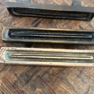 May include: Three rectangular bronze drawer pulls with a recessed, grooved design. The pulls have a weathered, antique appearance with variations in color, ranging from dark brown to a lighter, almost gold hue. The grooves run the length of the pulls.