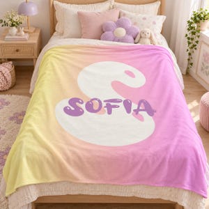 Puede incluir: Una manta de forro polar suave con un degradado de amarillo a rosa. En el centro, una gran forma blanca abstracta muestra el nombre SOFIA en letras moradas gruesas de estilo burbuja. La manta está extendida sobre una cama con almohadas.