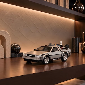 Puede incluir: Un coche modelo DeLorean plateado de máquina del tiempo con puertas de ala de gaviota, un conjunto de condensador de flujo trasero con cables rojos y una rejilla frontal negra con el logotipo DMC. Tiene ruedas detalladas.